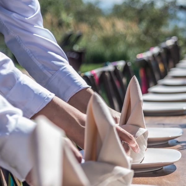 Hands in white shirts setting up an outdoor dining table with plates and folded napkins.
