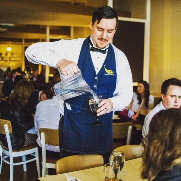 A Bangkok Garden waiter in a blue vest pours water for a customer in a busy dining room.