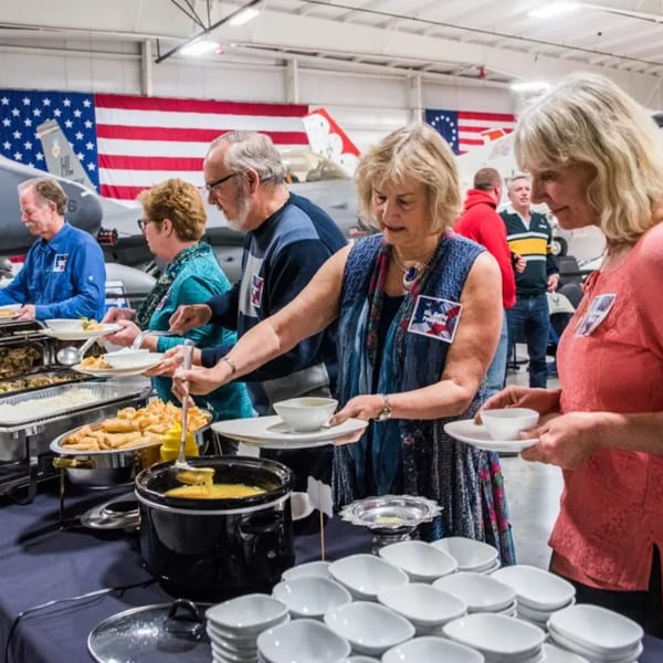 People serving themselves from a buffet line with Thai food, American flags, and aircraft in the background.