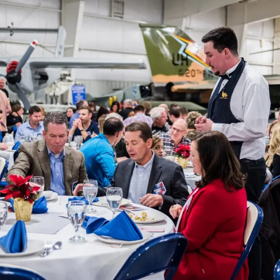 Diners seated at round tables with blue napkins and poinsettias, with airplanes in the background.
