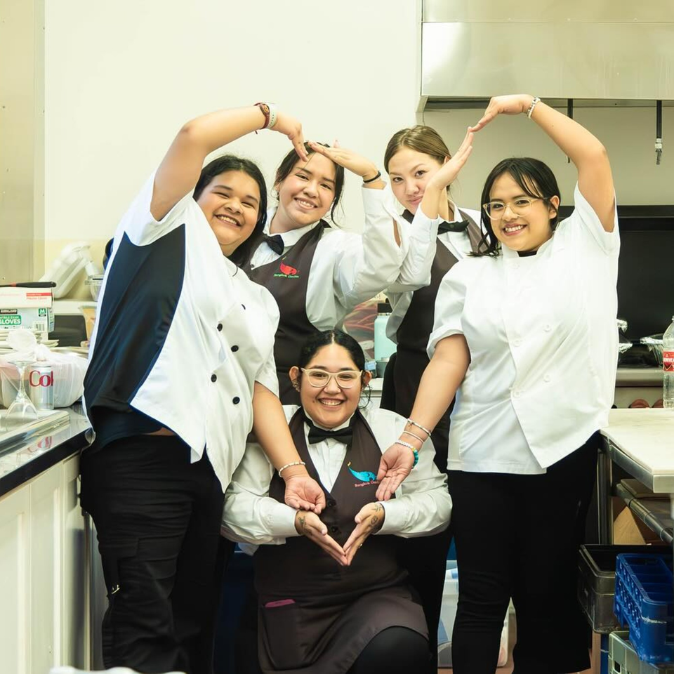 Five smiling Bangkok Garden staff members form heart shapes with their hands in a kitchen.