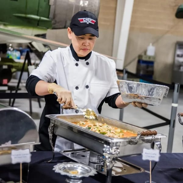 A chef in a black "CHEF" hat serves Thai red curry chicken from a buffet warmer.